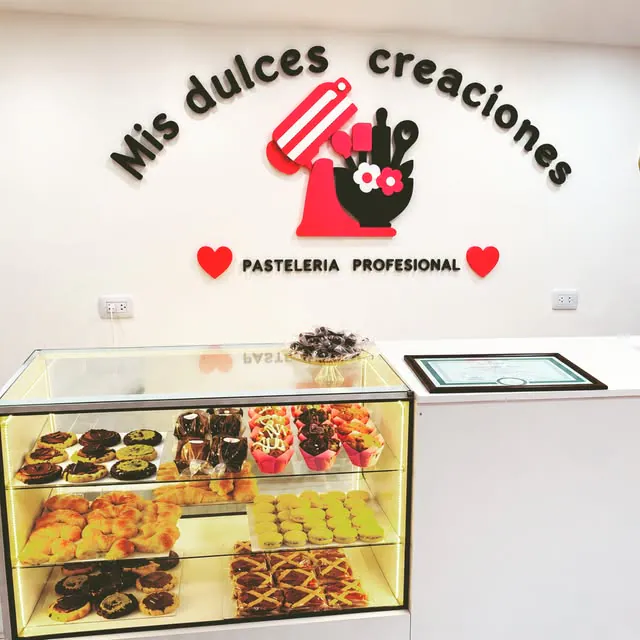 Pared blanca con el nombre y el logo de una pastelería en letras corpóreas negras y un mixer rojo y rosa, sobre una vitrina de pasteles